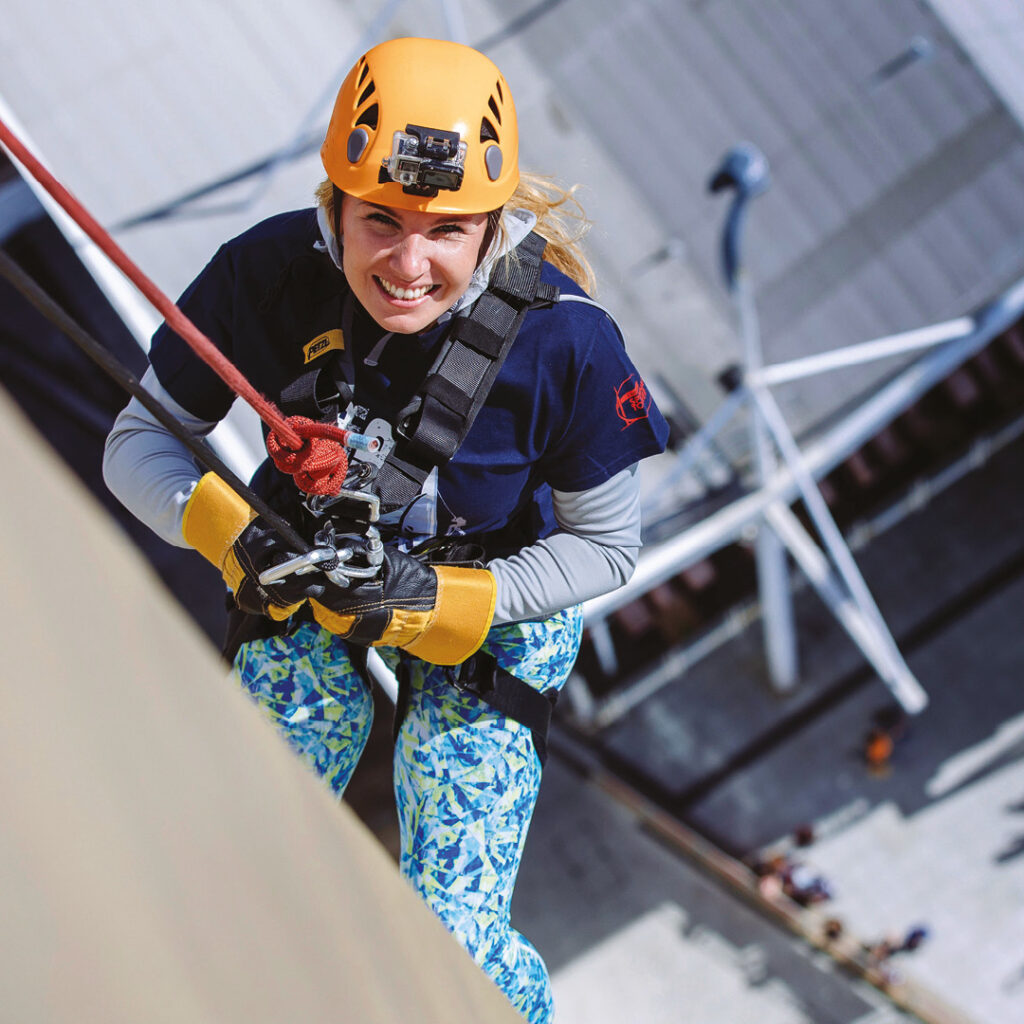 Person abseiling down the side of Spinnaker Tower and looking up to smile at the camera. 